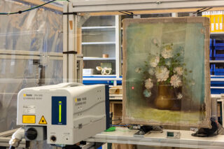 Ein wissenschaftlicher Scanner wird vor einem Blumenstillleben auf Leinwand positioniert, um es in einer Laborumgebung zu analysieren oder zu restaurieren.