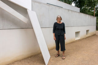 Eine Frau steht auf einem Kiesweg neben einem modernen Gebäude, an dessen Wand eine große weiße abstrakte Skulptur hängt.