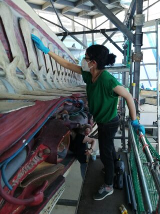 Eine Person mit Handschuhen und Maske steht auf einer Plattform und reinigt die Seite einer großen Skulptur in einem Industriegebiet.