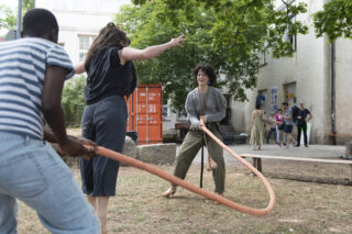 Drei Personen halten draußen einen großen Holzreifen und spielen damit, während andere im Hintergrund in der Nähe eines Gebäudes und eines orangefarbenen Containers stehen.