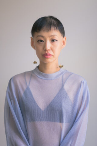 Eine Person mit kurzen dunklen Haaren, die ein durchsichtiges lavendelfarbenes Oberteil über einem dunklen Bralette und lange Ohrringe trägt und vor einem schlichten hellen Hintergrund steht.