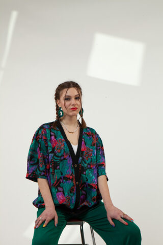 Eine Frau mit geflochtenen Haaren sitzt auf einem Hocker vor einem schlichten Hintergrund und trägt ein bunt gemustertes Hemd, grüne Hosen und Creolen.