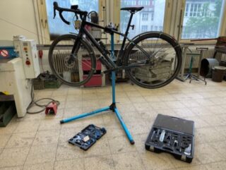 In einer Werkstatt ist ein Fahrrad auf einem Reparaturständer montiert, daneben stehen zwei offene Werkzeugkoffer auf dem Fliesenboden.