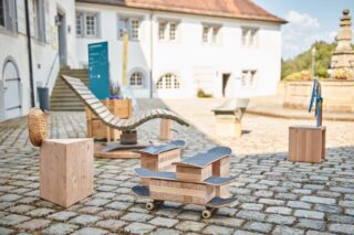 Ein Innenhof mit modernen Holzmöbeln, darunter ein Liegestuhl, Holzklötze und eine Bank aus Skateboards auf Rädern, auf einem Kopfsteinpflaster vor einem weißen Gebäude.