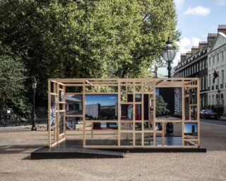 Im Freien sind in einer offenen Holzrahmenkonstruktion in ihren gitterartigen Abschnitten verschiedene Architekturfotografien zu sehen, im Hintergrund sind Bäume und Stadthäuser zu sehen.