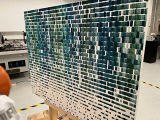 Eine Leinwand zeigt ein abstraktes Muster aus grünen und blauen rechteckigen Pinselstrichen in einem Raster, das auf einer Staffelei in einer Werkstatt steht.