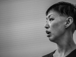 Schwarz-Weiß-Nahaufnahme einer Person mit kurzen Haaren, die nach rechts schaut, den Mund leicht geöffnet, vor einem unscharfen Hintergrund.