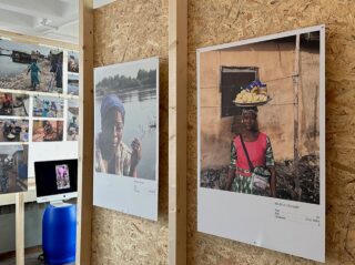 In einem Ausstellungsraum im Innenbereich sind an Holzwänden mehrere Dokumentarfotografien zu sehen. Im Hintergrund sind ein Computermonitor und ein blaues Fass zu sehen.