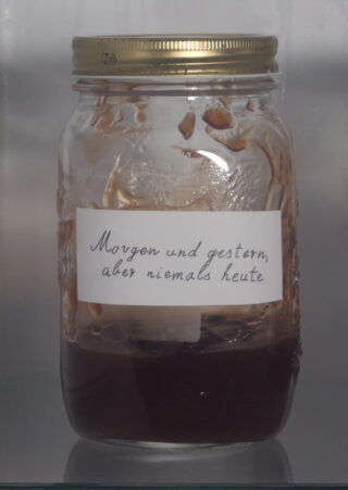 Ein Glasgefäß mit goldenem Deckel, teilweise gefüllt mit einer dunklen Substanz, mit der Aufschrift auf Deutsch: „Morgen und gestern, aber niemals heute.“.