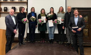 Eine Gruppe von Menschen steht auf einer Bühne; drei Frauen halten Blumen und Urkunden in den Händen, was auf eine Preisverleihung schließen lässt. Fünf Männer und Frauen stehen daneben, einige lächelnd, in einem formellen Innenraum.