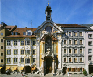 Barocke Kirchenfassade mit kunstvollen Verzierungen, Statuen und einem zentralen Bogeneingang, flankiert von Gebäuden mit detaillierten Fensterrahmen unter einem klaren blauen Himmel.