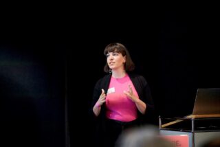 Eine Frau in einem rosa Hemd und einem schwarzen Blazer steht neben einem Laptop und gestikuliert mit ihren Händen, während sie eine Präsentation hält.