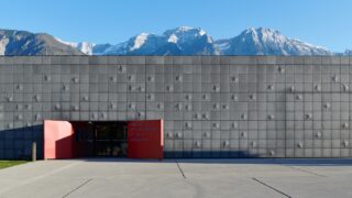 Ein modernes Gebäude mit strukturierter grauer Fassade und roten Eingangstüren vor der Kulisse schneebedeckter Berge unter einem klaren blauen Himmel.