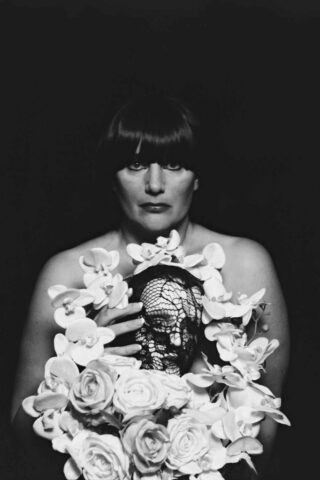 Eine Person mit dunklem Haar hält ein Blumenarrangement mit einem dekorativen Totenkopf und weißen Blumen vor einem dunklen Hintergrund.