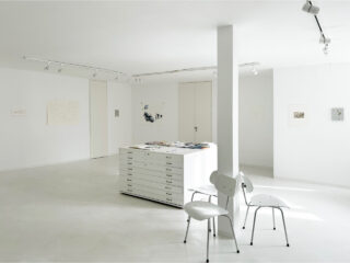 Minimalistische Kunstgalerie mit weißen Wänden, einem flachen Aktenschrank mit Papieren, drei weißen Stühlen und mehreren an den Wänden hängenden Kunstwerken.