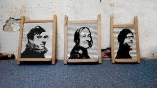 Drei alte Waschbretter mit schwarz-weiß gemalten Gesichtsporträts, lehnen an einer abgenutzten, weißen Wand auf einem blauen Teppichboden.