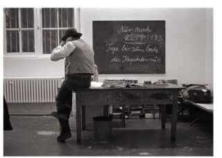 Eine Person sitzt auf der Kante eines Tisches in einem Klassenzimmer. Hinter ihr befindet sich eine Tafel mit deutschen Texten und Zahlen und auf der linken Seite ein Fenster.