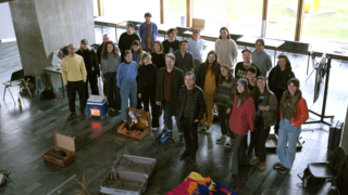 Eine Gruppe von etwa 25 Personen steht im Innenbereich auf einem Steinboden, umgeben von offenen Koffern und Geräten, im Hintergrund große Fenster.