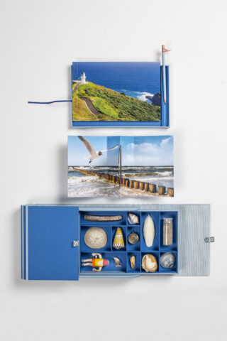 Eine blaue Schachtel mit Fächern, in denen sich Muscheln und kleine Gegenstände befinden, wird neben zwei offenen Fotobüchern mit Küsten- und Meeresszenen ausgestellt.