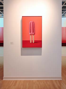 An einer weißen Galeriewand hängt ein gerahmtes Gemälde eines rosa Eis am Stiel mit zwei Stielen, beleuchtet von einer Deckenbeleuchtung.