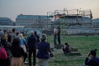 Eine kleine Menschenmenge versammelt sich im Freien, um zwei Personen zuzuhören, die auf einer erhöhten Plattform sprechen. Im Hintergrund sind mit Graffiti bedeckte Wände und ein Gerüst zu sehen.