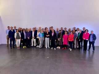 Eine Gruppe von etwa vierzig Erwachsenen steht zusammen in einem Innenbereich vor einer schlichten hellvioletten Wand und posiert für ein Gruppenfoto.