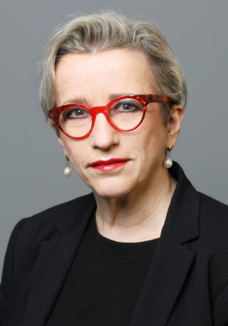 Eine Frau mit kurzen grauen Haaren, einer roten Brille, Perlenohrringen, rotem Lippenstift und einem schwarzen Blazer posiert vor einem schlichten grauen Hintergrund.