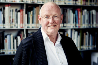 Ein Mann mit Brille und weißem Hemd steht vor mit Büchern gefüllten Bücherregalen und lächelt in die Kamera.