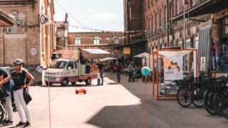 Menschen versammeln sich in einem städtischen Innenhof mit Imbisswagen, Fahrrädern und Vordächern; an einem sonnigen Tag hängen bunte Wimpel über ihnen.