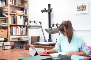 Eine Frau sitzt an einem Schreibtisch in einer Bibliothek oder einem Büro, arbeitet an einem Tablet und liest Dokumente, im Hintergrund sind Bücherregale und Scan-Geräte zu sehen.