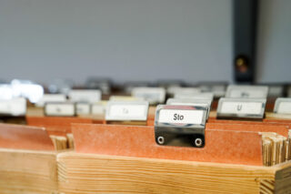 Eine Reihe von Aktenordnern mit beschrifteten Registern, darunter einer mit der Aufschrift „Sto“, angeordnet in einer hölzernen Aktenschublade.