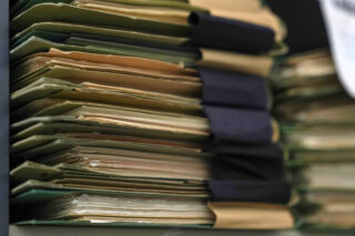 Eine Nahaufnahme mehrerer Stapel Papierakten, die mit großen schwarzen Heftklammern zusammengehalten und auf einem Regal angeordnet sind.