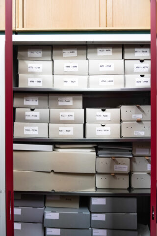 Ein Metallschrank mit beschrifteten Archivboxen, die auf Regalen angeordnet sind und auf der Vorderseite jeder Box eine sichtbare Nummerierung aufweisen.