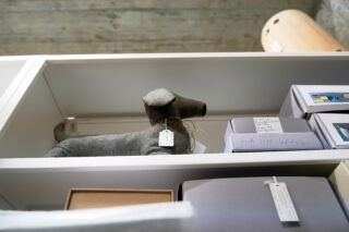 Ein grauer Türstopper aus Stoff in Hundeform mit einem Etikett steht auf einem weißen Regal neben verpackten Artikeln in einem Geschäft.