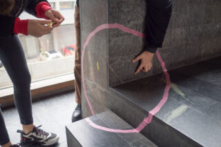 Zwei Personen zeichnen mit rosa Klebeband einen Kreis an der Kante einer Steintreppe entlang, wobei eine Person zeigt und die andere das Klebeband hält.