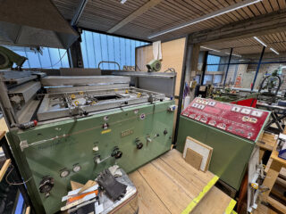 Eine große Industriemaschine mit Bedienfeldern, Messgeräten und Schaltern steht auf einer Holzplattform in einer mit Geräten gefüllten Werkstatt.