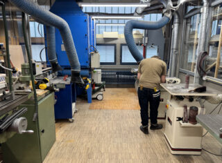Eine Person steht bei der Arbeit in einer Werkstatt voller Industriemaschinen, Lüftungsrohre und Holzbearbeitungswerkzeuge.