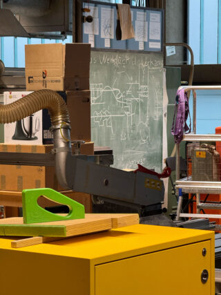 Eine Werkstatt mit gestapelten Kisten, einem grünen Elektrowerkzeug auf einem gelben Tisch, einer Tafel mit Diagrammen und einem metallenen Lüftungsschacht im Hintergrund.