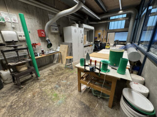 Eine Werkstatt mit Industrieausrüstung, Werkbänken, Kunststoffrohren, Werkzeugen, Lagerschränken und verschiedenen Behältern, die im Raum mit Betonboden verstreut sind.