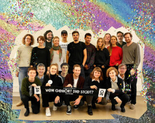 Eine Gruppe von achtzehn Personen posiert im Innenbereich vor einer mit buntem Konfetti und gemalten Mustern dekorierten Wand; einige halten ein Banner mit der Aufschrift „Wem gehört die Stadt?“.