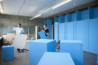 Zwei Personen in einem Raum bauen blaue Schränke zusammen, wobei eine Person zeigt und die andere eine Platte hält; im Hintergrund ist eine grüne Tafel zu sehen.