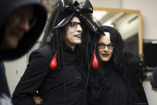Zwei Personen in schwarzen Outfits im Gothic-Stil und mit Perücken stehen zusammen im Innenbereich; eine trägt eine große schwarze Kopfbedeckung mit Schleife und roten Tropfenornamenten, beide tragen eine Brille.