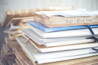 Ein Stapel Ordner, Dokumente und Pakete, eines davon mit der Aufschrift „Hier öffnen bitte“ ganz oben, auf einer flachen Fläche angeordnet.