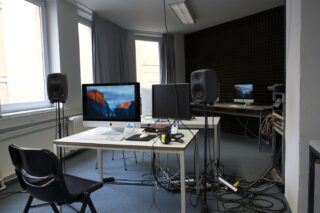 Moderner Büro- oder Studioraum mit Schreibtischen, Computern, mehreren Monitoren, Lautsprechern und sichtbaren Kabeln auf dem Boden; im Hintergrund ist eine dunkle, schalldichte Wand.