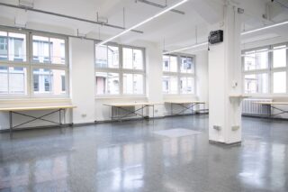 Ein heller, leerer Raum mit großen Fenstern, weißen Wänden, Leuchtstoffröhren, poliertem grauen Boden und mehreren langen Schreibtischen an den Wänden.