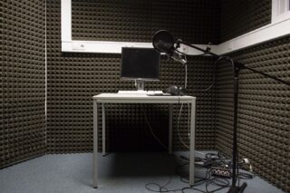 Ein kleines Aufnahmestudio mit schalldichten Schaumstoffwänden, einem Schreibtisch mit Monitor, Tastatur und Maus sowie einem Mikrofon auf einem Ständer mit Kabeln auf dem Boden.