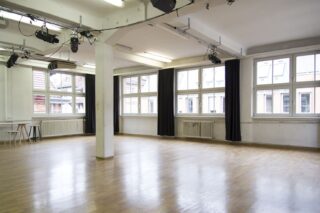 Geräumiger, leerer, gut beleuchteter Studioraum mit Holzböden, großen Fenstern, schwarzen Vorhängen, Heizkörpern und an der Decke montierter Beleuchtungsanlage.