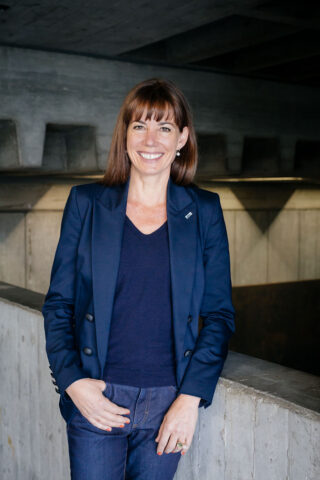 Eine Frau mit glattem braunem Haar, die einen marineblauen Blazer und Jeans trägt, steht in einem Gebäude vor einer Betonwand und lächelt in die Kamera.