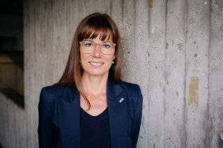 Eine Frau mit langen braunen Haaren und Brille sowie einem dunklen Blazer steht vor einer strukturierten Betonwand und lächelt in die Kamera.