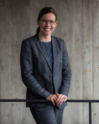Eine Frau mit Brille und dunklem Anzug steht in einem Innenbereich vor einer Betonwand und lächelt in die Kamera, während ihre Hände auf einem Geländer ruhen.